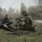 Três personagens de Escape from Tarkov em campo aberto, com colete tático, uniforme de camuflagem, armas e mochila militar no gramado